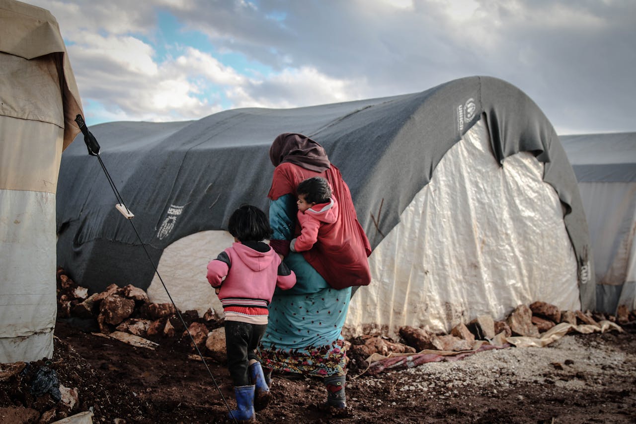 about-us A woman and children walk near tents in a refugee camp in Idlib, showcasing resilience amidst adversity.