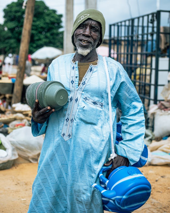services-02 An elderly man dressed in traditional attire holding household items at an outdoor market, smiling warmly.